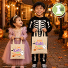 A young child in a skeleton costume and a toddler in a lavender princess dress stand outdoors at night holding matching tote bags printed with the phrase 'Too Cute to Spook' and Halloween graphics. The scene is warmly lit by string lights and pumpkins, and a circular badge in the top right says the file includes one design and cut files/sublimation.