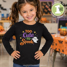A young girl with brown pigtails smiles while wearing a black long-sleeve shirt with a colorful 'Too Cute to Spook' Halloween graphic. The graphic features orange text, a jack-o'-lantern, purple text, yellow stars, and a white ghost.