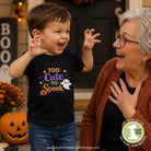 A young boy wearing a black graphic t-shirt that reads 'Too Cute to Spook' in purple, orange, and white, standing next to an older woman in a cozy outdoor setting with pumpkins and fall decorations.