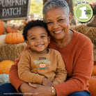 A smiling older woman and young boy sit together in a pumpkin patch setting. The boy wears a tan long-sleeve shirt with a 'Cuteest Pumpkin in the Patch' graphic, and the woman wears an rust-orange knit sweater.