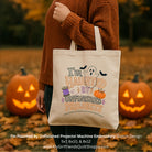 A person in a rust-brown sweater holds a cream canvas tote bag outdoors with jack-o'-lanterns in the background. The tote bag has a machine-embroidered Halloween design that reads 'I'm Haunted by Unfinished Projects' with a ghost, bats, pumpkin, and purple potion bottle.