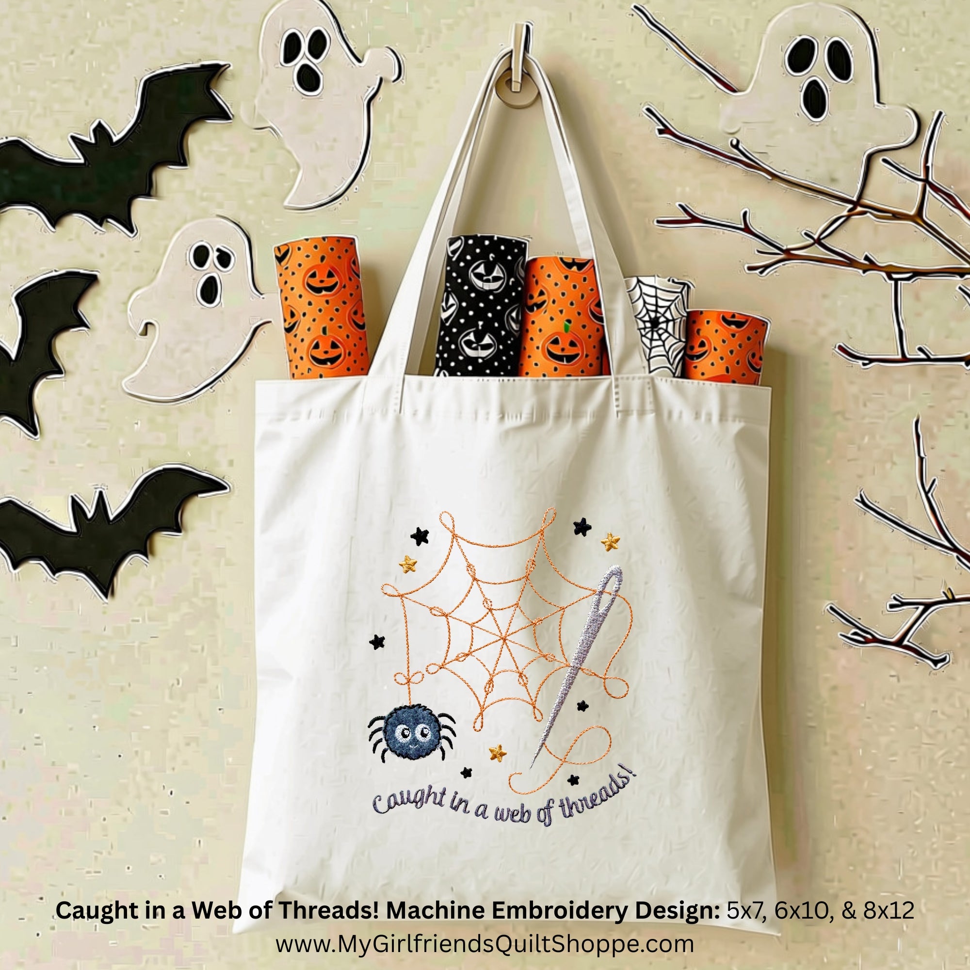 A cream canvas tote bag with a Halloween-themed machine embroidery design showing an orange spiderweb, a blue spider, stars, and the text 'Caught in a web of threads!'. The bag is shown filled with patterned fabric rolls, surrounded by Halloween decorations like ghosts, bats, and branches on a textured beige background.