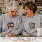 Two women smiling and laughing while wearing matching light gray crewneck sweatshirts with a colorful embroidered design that reads 'My Broomstick Runs on Bobbin Thread.' A sewing machine and quilting supplies are visible on a table in a home-style craft setting.