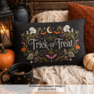 A square black decorative pillow with embroidered 'Trick-or-Treat' text and autumn motifs, styled on a cozy knit blanket with pumpkins, a mug, and pillows in a fall-themed setting.