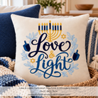 A square throw pillow shown in a cozy living-room setting, featuring a Hanukkah-themed machine embroidery design with the words “Love & Light” in dark blue script, a gold menorah, blue dreidels, blue floral accents, and gold Star of David motifs. Text at the bottom reads “Love & Light Hanukkah Machine Embroidery Design 6x10, 8x12, 9x14” and includes the website MyGirlfriendsQuiltShoppe.