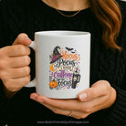 A person holds a white ceramic mug with a colorful Halloween-themed graphic that reads 'Hocus Pocus I Need Caffeine to Focus' with a witch hat, pumpkin, spider, and coffee cup illustration. The person is wearing a black knit sweater, and the mug is shown against a dark background.