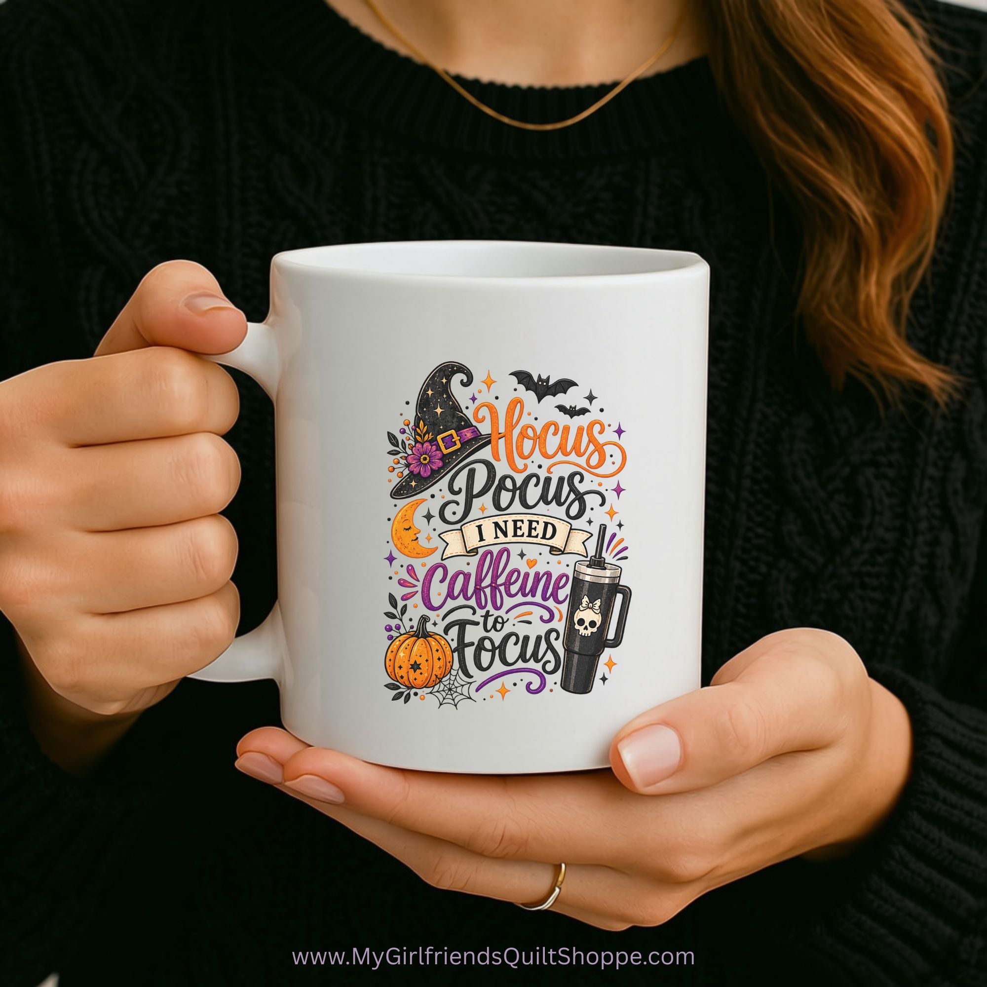 A person holds a white ceramic mug with a colorful Halloween-themed graphic that reads 'Hocus Pocus I Need Caffeine to Focus' with a witch hat, pumpkin, spider, and coffee cup illustration. The person is wearing a black knit sweater, and the mug is shown against a dark background.