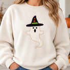A woman wearing a cream-colored crewneck sweatshirt with a graphic of a ghost wearing a black witch hat with orange stars and a green band, paired with blue jeans.