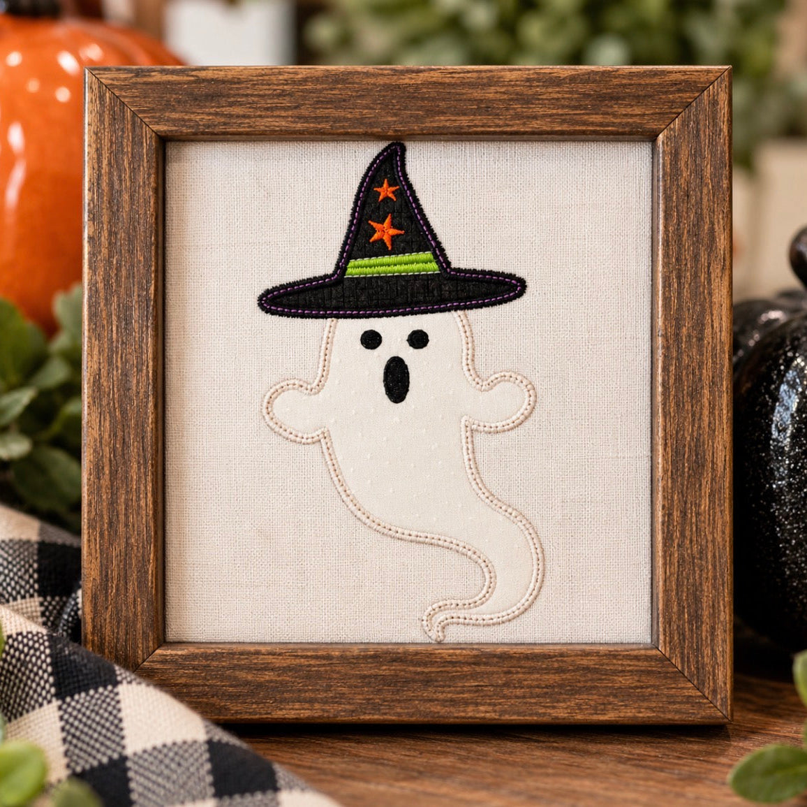 A framed Halloween decorative piece featuring an embroidered white ghost wearing a black witch hat with orange stars and a green band, set in a rustic brown wooden frame on a tabletop with seasonal decor in the background.