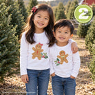 Two children standing outdoors in a Christmas tree lot wearing matching white long-sleeve tops with gingerbread man graphics. A green circular badge in the top right says it includes two designs, and a website watermark appears at the bottom left.
