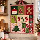 A festive Christmas quilt hanging on a wall in a decorated kitchen. The quilt features patchwork panels with gingerbread men, candy canes, stockings, snowflakes, a Christmas tree, and 'HO HO' motifs in red, green, beige, and white.