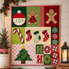 A festive quilted wall hanging or blanket featuring Christmas-themed patchwork panels with gingerbread men, candy canes, stockings, snowflakes, a Christmas tree, and the word HOHO. The quilt is displayed against a white shiplap wall with holiday greenery and a lit lantern nearby.