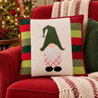 A square Christmas throw pillow with a gnomes face design, styled on a red armchair next to a green knit pillow and a cozy knit blanket, with a decorated Christmas tree in the background.