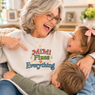 A woman and two children are sitting together on a couch, smiling and laughing. The woman wears a light beige crewneck sweatshirt with a colorful graphic that reads 'MiMi Fixes Everything' in red, green, yellow, and blue lettering.