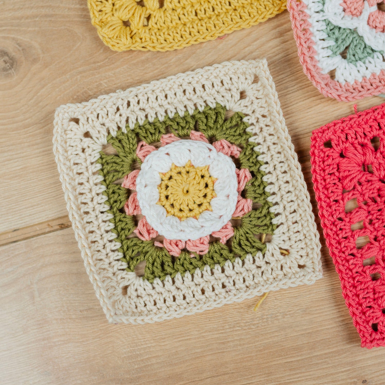 A close-up of a handmade crocheted granny square in cream, olive green, white, and pink, laid flat on a light wood surface. Other colorful crocheted squares are partially visible in the corners.