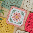 A flat-lay arrangement of colorful handmade crocheted granny squares on a light wood background, featuring a central pink and white floral square surrounded by other multi-colored squares.