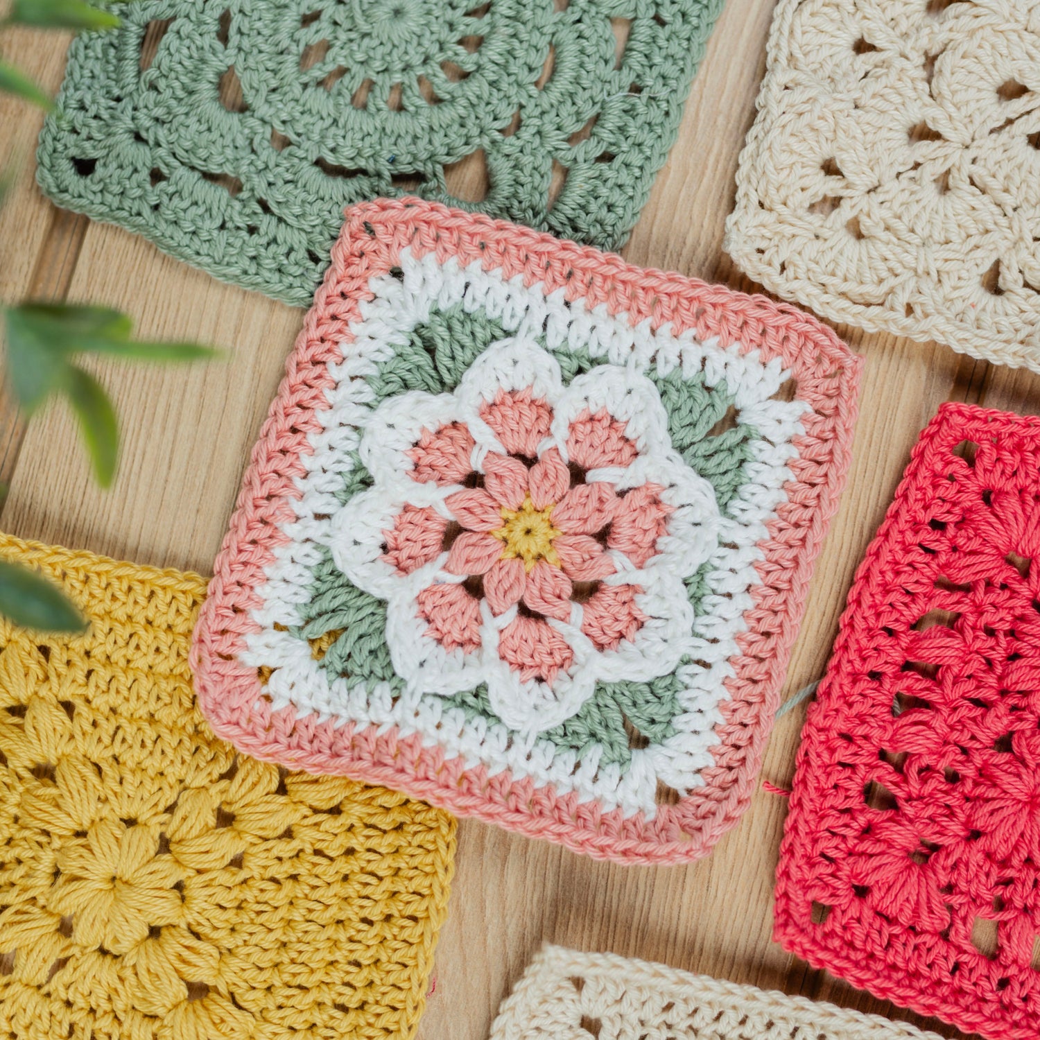 A flat-lay arrangement of colorful handmade crocheted granny squares on a light wood background, featuring a central pink and white floral square surrounded by other multi-colored squares.