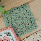 A handmade crocheted granny square in sage green yarn, laid flat on a light wood surface next to other colorful crochet squares.