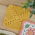 A yellow crocheted square dishcloth or coaster with an intricate floral pattern, displayed on a light wood tabletop next to other colorful crocheted pieces and a green plant.