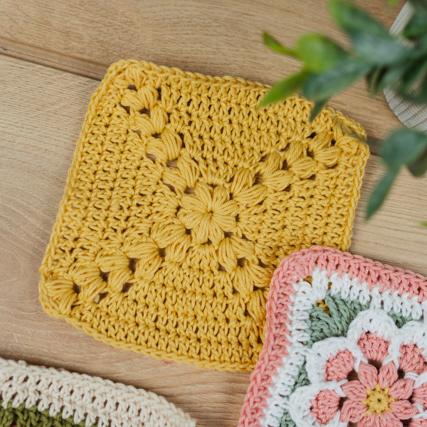 A yellow crocheted square dishcloth or coaster with an intricate floral pattern, displayed on a light wood tabletop next to other colorful crocheted pieces and a green plant.