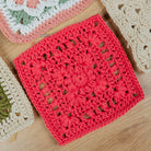 A bright red crocheted granny square with a floral lace pattern, displayed on a light wood surface alongside other colorful crochet squares.