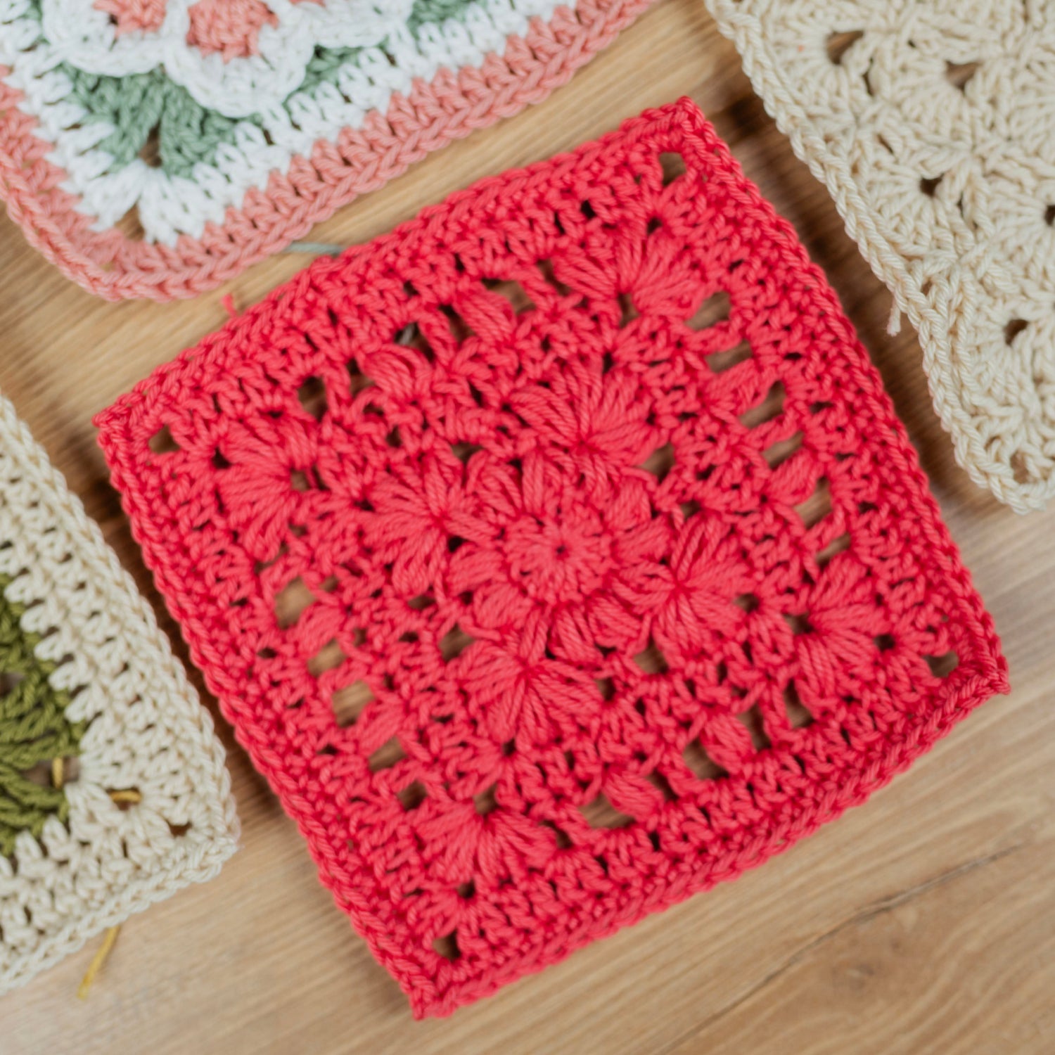 A bright red crocheted granny square with a floral lace pattern, displayed on a light wood surface alongside other colorful crochet squares.