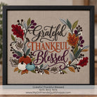 Framed cross-stitch artwork with the words “Grateful Thankful Blessed” surrounded by autumn leaves, berries, and small pumpkins on a beige fabric background, shown in a black frame against a softly blurred indoor backdrop.