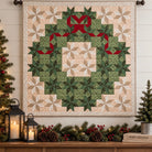 A large quilted wall hanging with a Christmas wreath design, featuring green and beige geometric patterns with a red ribbon bow, displayed in a decorated holiday room setting.