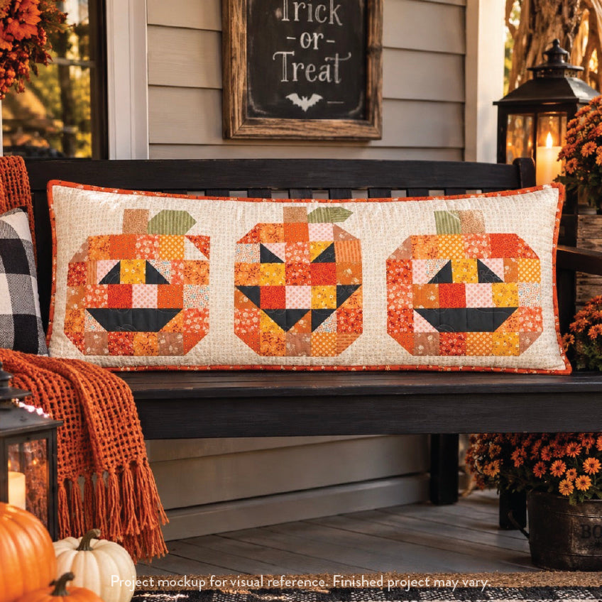 A rectangular patchwork pumpkin-themed porch pillow displayed on a dark wooden bench in a fall porch setting. The pillow features three stylized jack-o'-lantern faces made from orange, yellow, and floral patterned fabric blocks on an off-white background.
