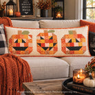 A rectangular quilted throw pillow with three stitched jack-o'-lantern faces in orange, yellow, and black on a cream background, styled on a beige sofa in a cozy living room setting.