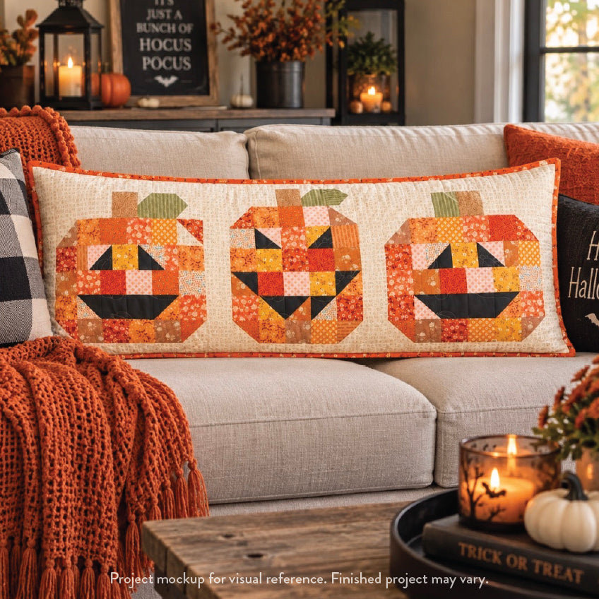 A rectangular quilted throw pillow with three stitched jack-o'-lantern faces in orange, yellow, and black on a cream background, styled on a beige sofa in a cozy living room setting.