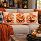 A rectangular patchwork pumpkin bench pillow with three carved jack-o'-lantern faces sits on a dark wooden porch bench. The pillow features a cream background with orange, yellow, black, and green patchwork pumpkin designs.