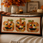 A rectangular decorative throw pillow with three patchwork jack-o'-lantern faces sits on a cream couch in a cozy autumn living room. The pillow has a black border and is styled with a chunky knit blanket, with fall decorations like pumpkins, a vase of leaves, and a 'Hello fall' sign in the background.