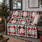 A quilted throw blanket with a rustic Christmas star pattern in red, green, and gray is draped over a weathered wooden bench on a porch. The background features a decorated Christmas tree, a framed sign reading 'good neighbors warm hearts', and holiday pillows.