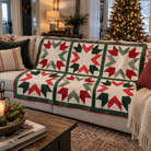 A Christmas-themed quilted throw blanket draped over a light beige sofa in a cozy living room. The blanket features a classic starburst pattern in red, dark green, and cream, with a solid dark green border.