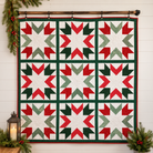 A large quilted wall hanging with a Christmas star pattern, featuring red, green, and white blocks, hung on a wooden rod against a white shiplap wall with holiday greenery and a lantern.