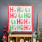 A Christmas-themed quilt hanging on a gold rod against a dark gray wall, featuring the word 'HO HO HO' in red and green with small tree and cabin motifs. The quilt is displayed in a styled room setting above a wooden console table with plants and decor.