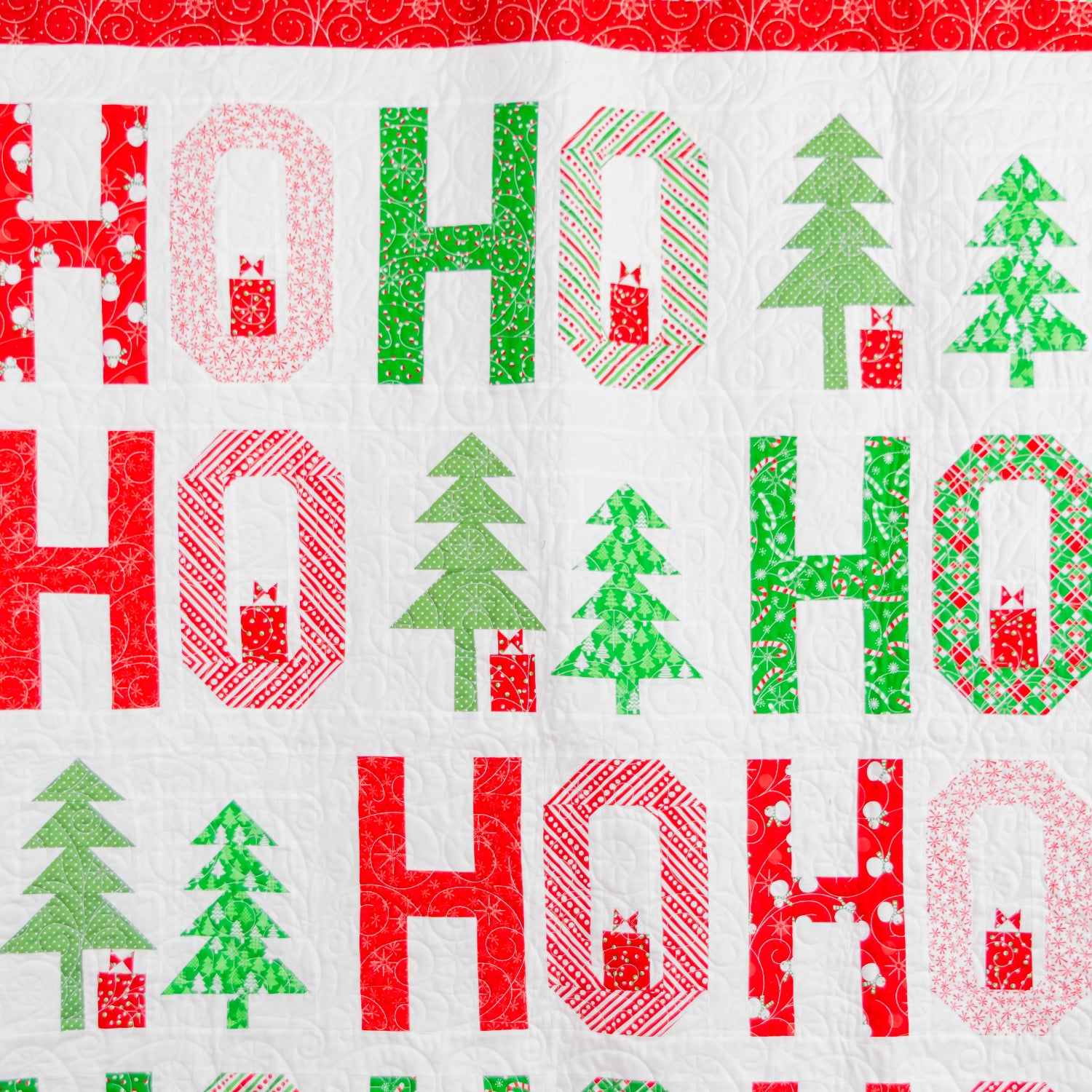 A close-up of a quilted fabric featuring a festive 'HO HO HO' holiday pattern with red and green text and Christmas tree motifs on a white background.