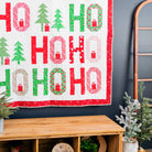 A festive quilted wall hanging with the word 'HO HO HO' in large red and green letters, decorated with Christmas trees and snowflake motifs, displayed on a dark wall above a wooden bench with holiday plants.
