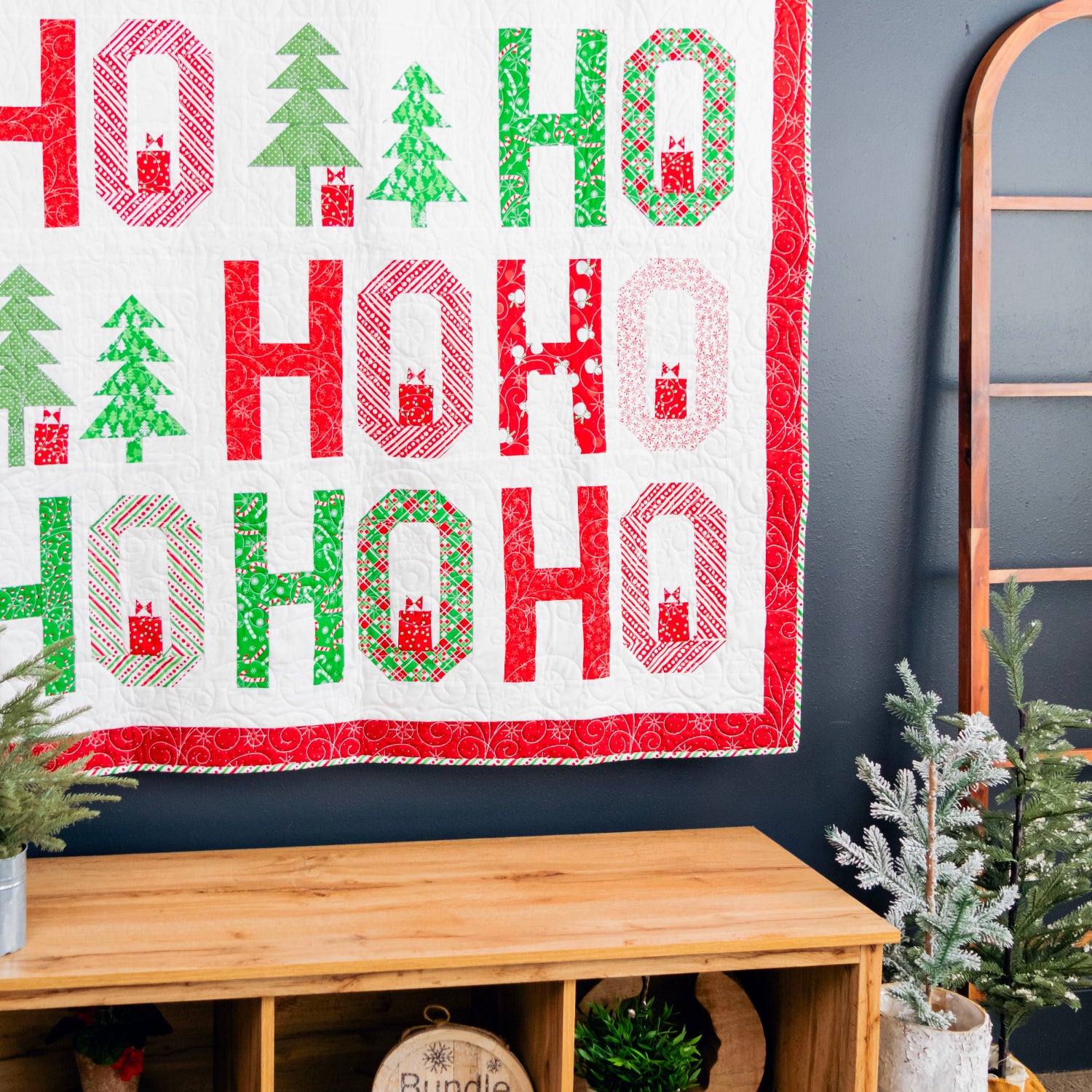 A festive quilted wall hanging with the word 'HO HO HO' in large red and green letters, decorated with Christmas trees and snowflake motifs, displayed on a dark wall above a wooden bench with holiday plants.
