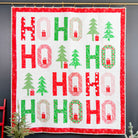 A handmade Christmas quilt hanging on a dark gray wall, featuring a white background with red and green 'HO HO HO' letters, green Christmas trees, and red gingerbread houses, framed by a red patterned border.
