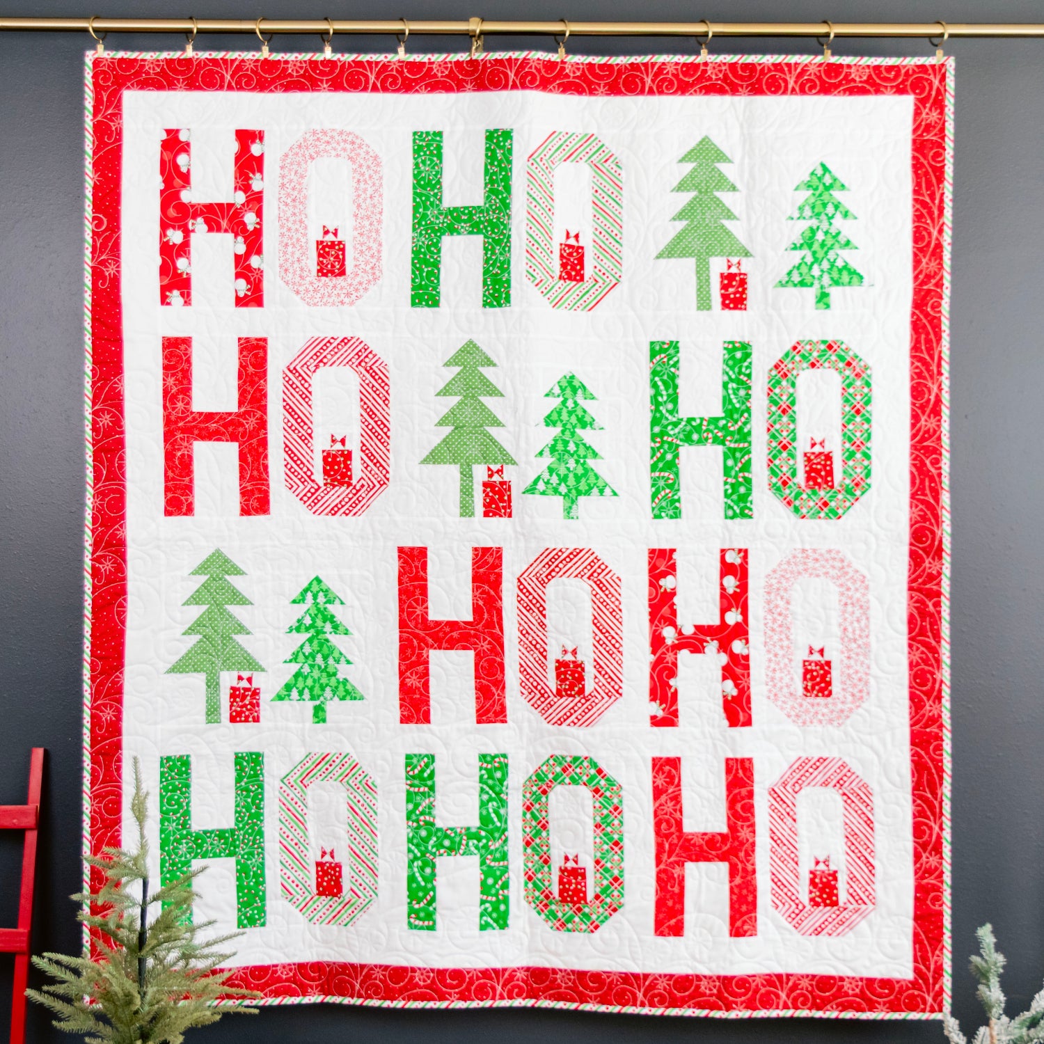 A handmade Christmas quilt hanging on a dark gray wall, featuring a white background with red and green 'HO HO HO' letters, green Christmas trees, and red gingerbread houses, framed by a red patterned border.