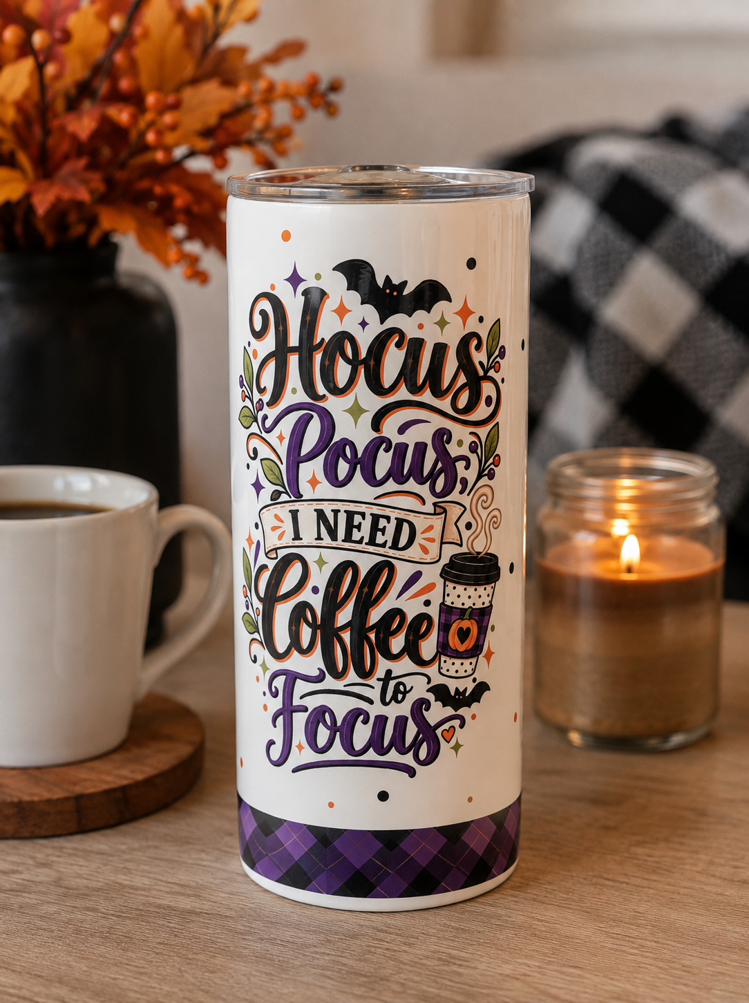 A white insulated tumbler with a clear lid sits on a wooden table, surrounded by a white coffee mug, a lit candle in a glass jar, and autumn decor. The tumbler features the phrase 'Hocus Pocus, I Need Coffee to Focus' in stylized lettering with bats, stars, and a purple plaid band at the bottom.