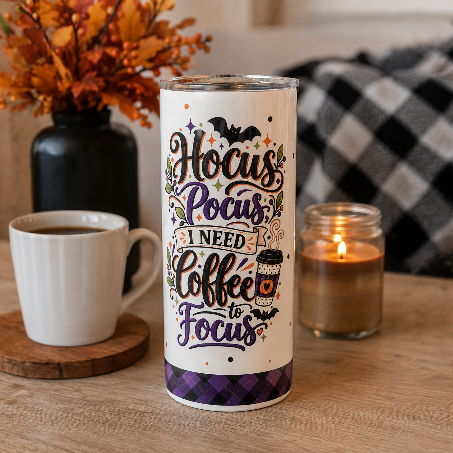 A tall insulated tumbler with a Halloween-themed quote reading 'Hocus Pocus I Need Coffee to Focus' sits on a wooden table. The tumbler is styled with a white coffee mug, a lit candle, and autumn decorations in the background.