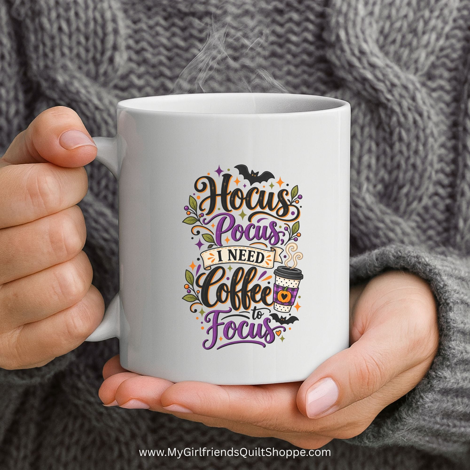 A person holds a white ceramic mug with a colorful graphic that reads 'Hocus Pocus I Need Coffee to Focus' with bats, flowers, and a coffee cup illustration. The person is wearing a gray cable-knit sweater, and steam is visible at the top of the mug.