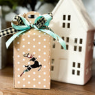 Gift box with polka dots and reindeer design, tied with a green ribbon, in front of a ceramic house decoration.