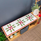 A Christmas-themed quilted bench runner with three green and red snowflake patterns on a white background, styled on a wooden bench in a decorated room.