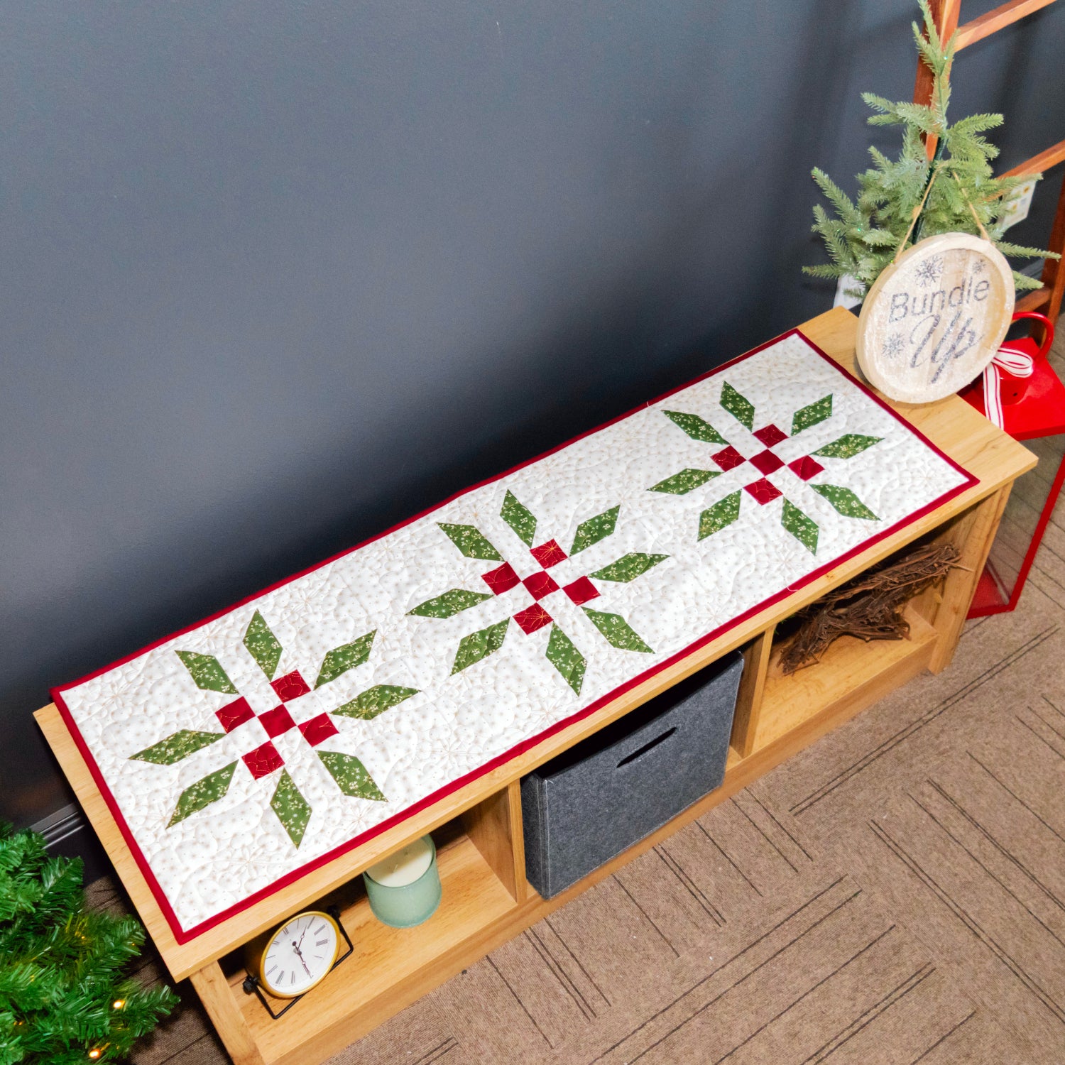 A Christmas-themed quilted bench runner with three green and red snowflake patterns on a white background, styled on a wooden bench in a decorated room.