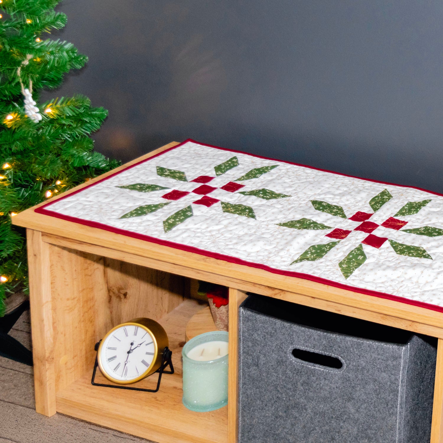A long quilted table runner with a Christmas starburst pattern in green and red on a white background, displayed on a wooden console table in a decorated holiday setting.