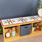 A wooden bench with open storage compartments, decorated with a white quilted holiday table runner featuring green pine branches and red bows. The bench is styled in a cozy room setting with a Christmas tree, a gray wall, and a ladder shelf in the background.